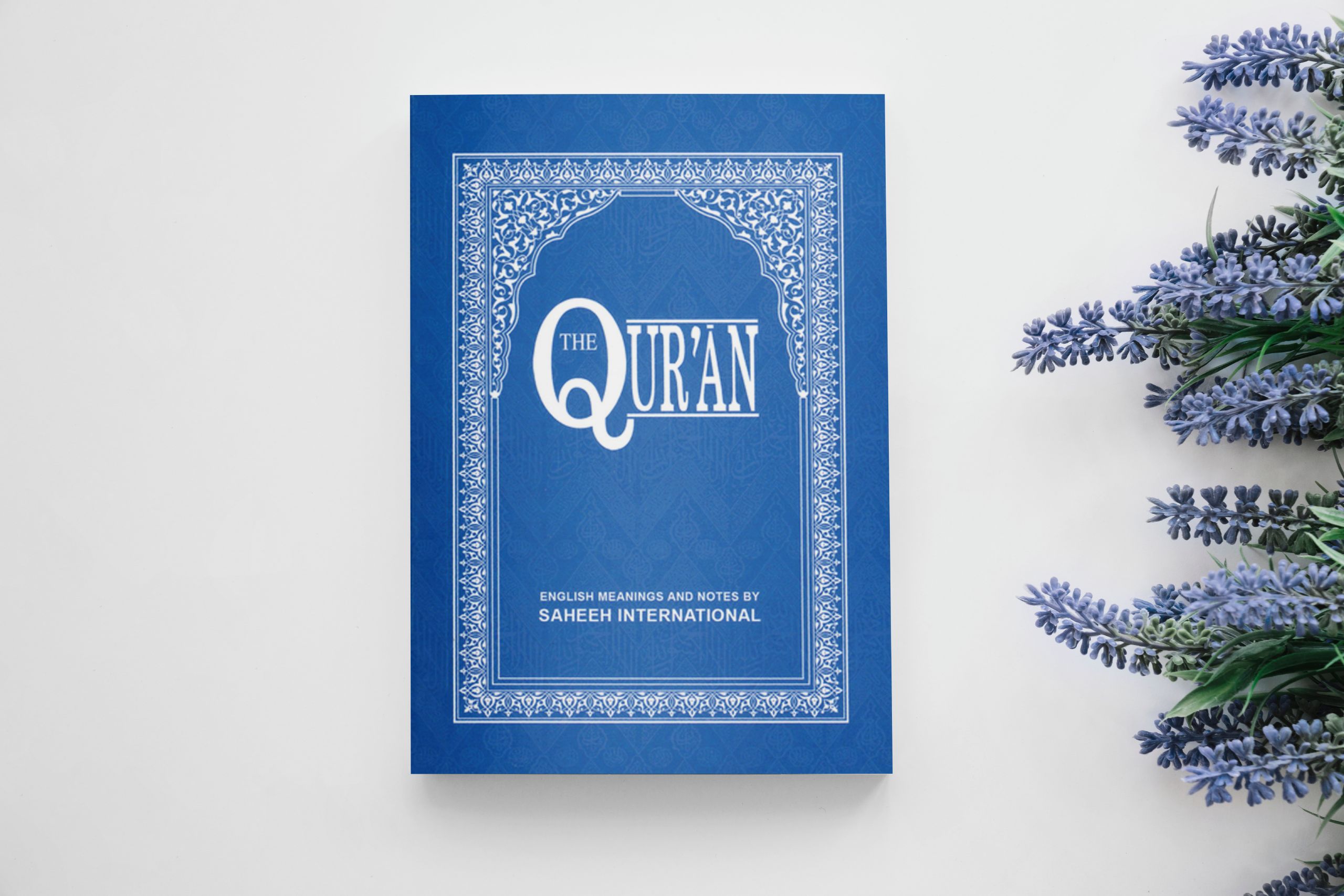 A paperback printed copy of an English translation of the Quran. The cover is blue and has an arch-like design as the border. The text on the cover reads: "The Qur'an: English Translation of the Meanings by Saheeh International"